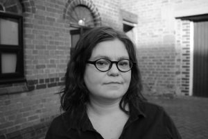 A black and white close-up photograph of author Abigail Ryan who is looking directly into the camera with brick buildings in the background.